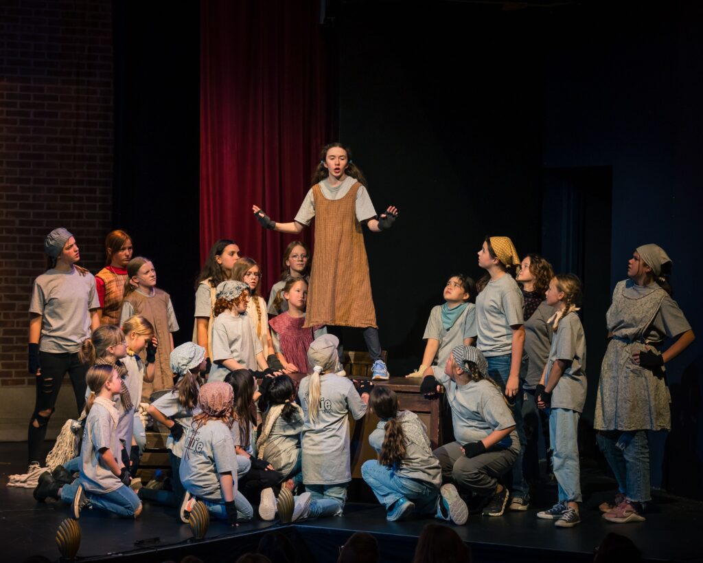 Group of young performers performing Annie. Annie is standing on a box, while the other orphans look on.